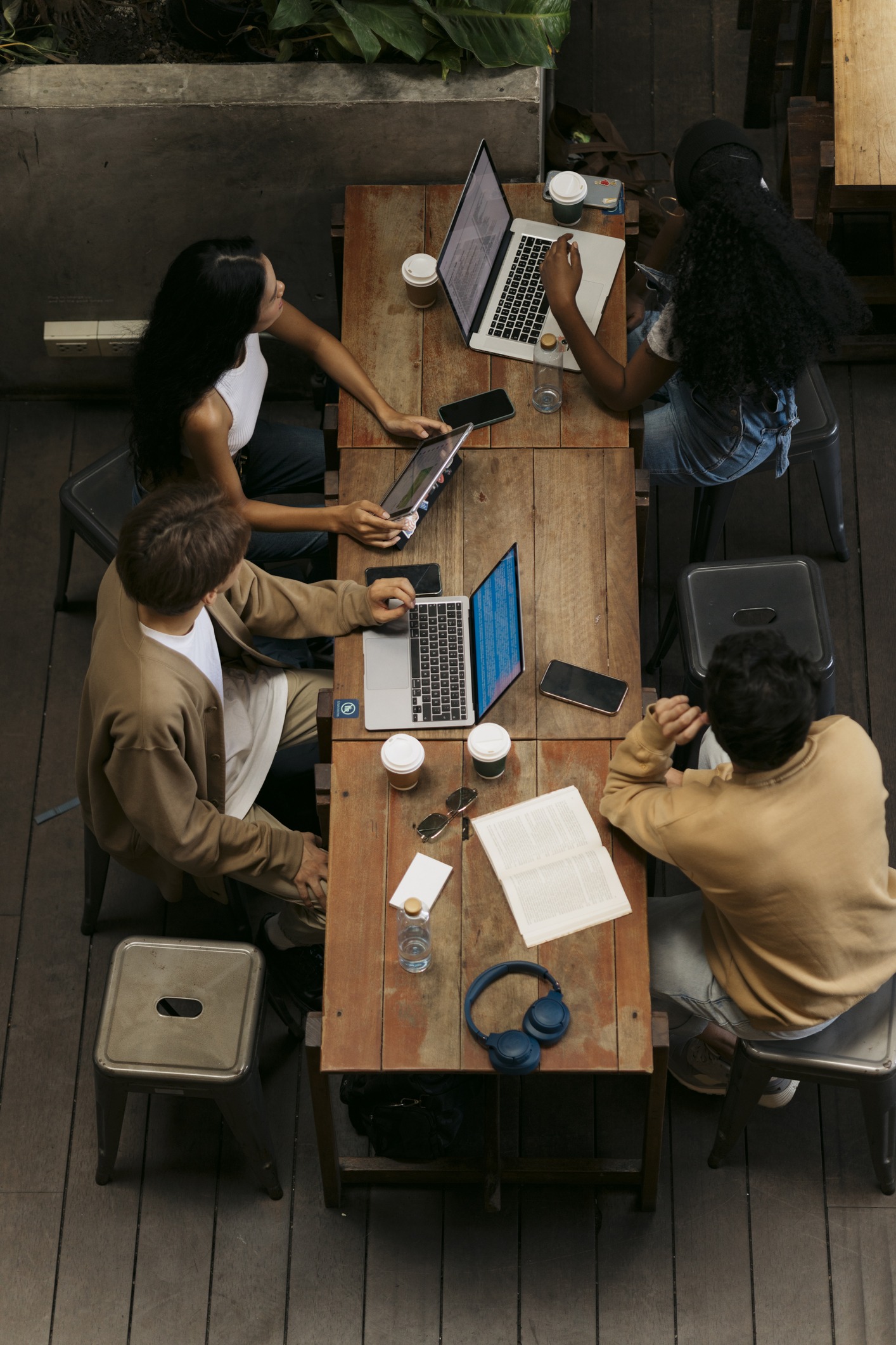Top view of young adult students studying together with laptop and digital tablet in a coffee shop and looking away. Group of diverse young people studying in coffee house together.