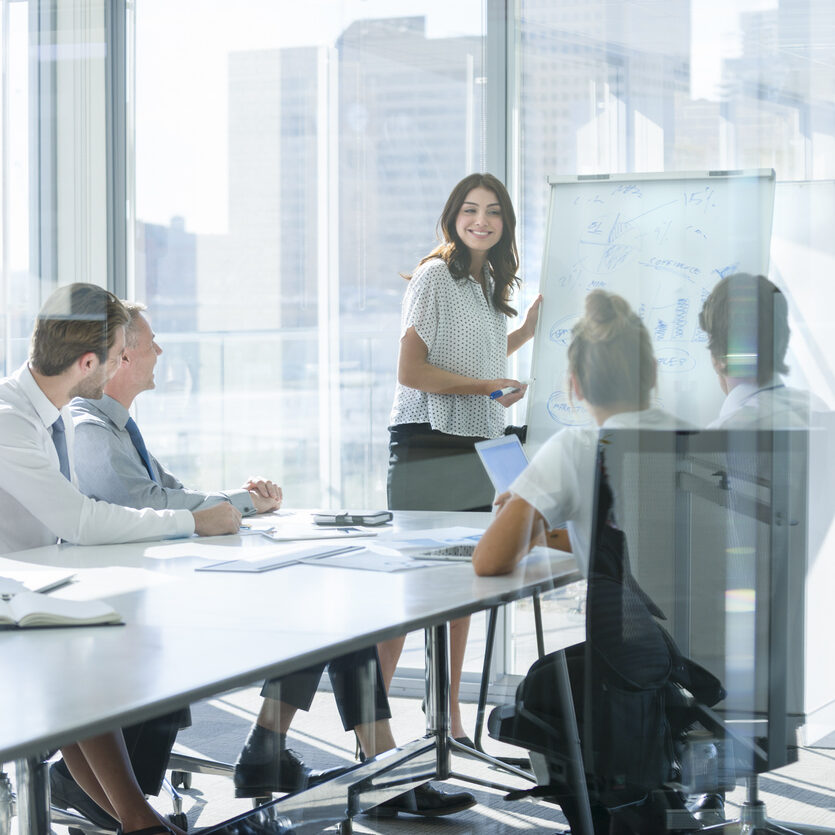 A smiling woman presents charts on a whiteboard to a diverse team in an office boardroom.