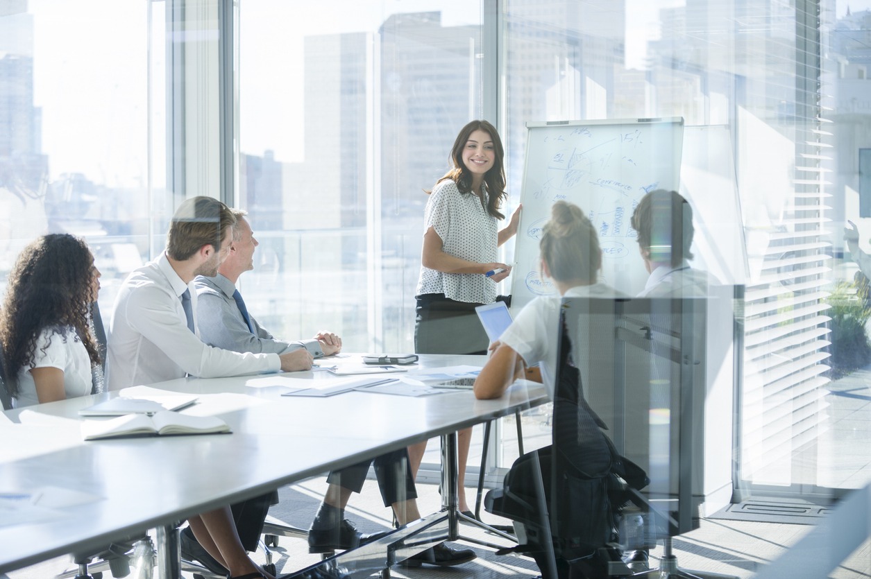 A smiling woman presents charts on a whiteboard to a diverse team in an office boardroom.